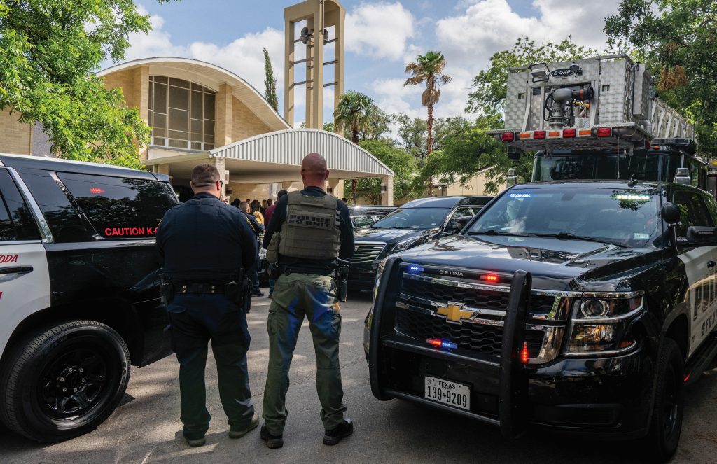 Two officers near police and fire vehicles outside church with tall bell tower; trees and parked cars surround the scene.