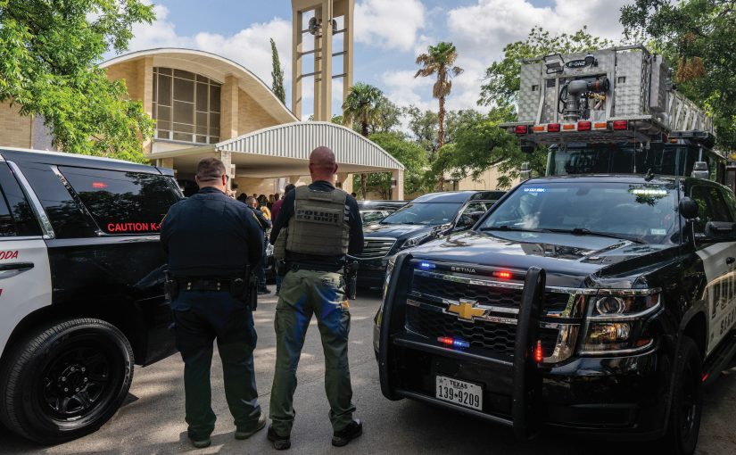 Two officers near police and fire vehicles outside church with tall bell tower; trees and parked cars surround the scene.