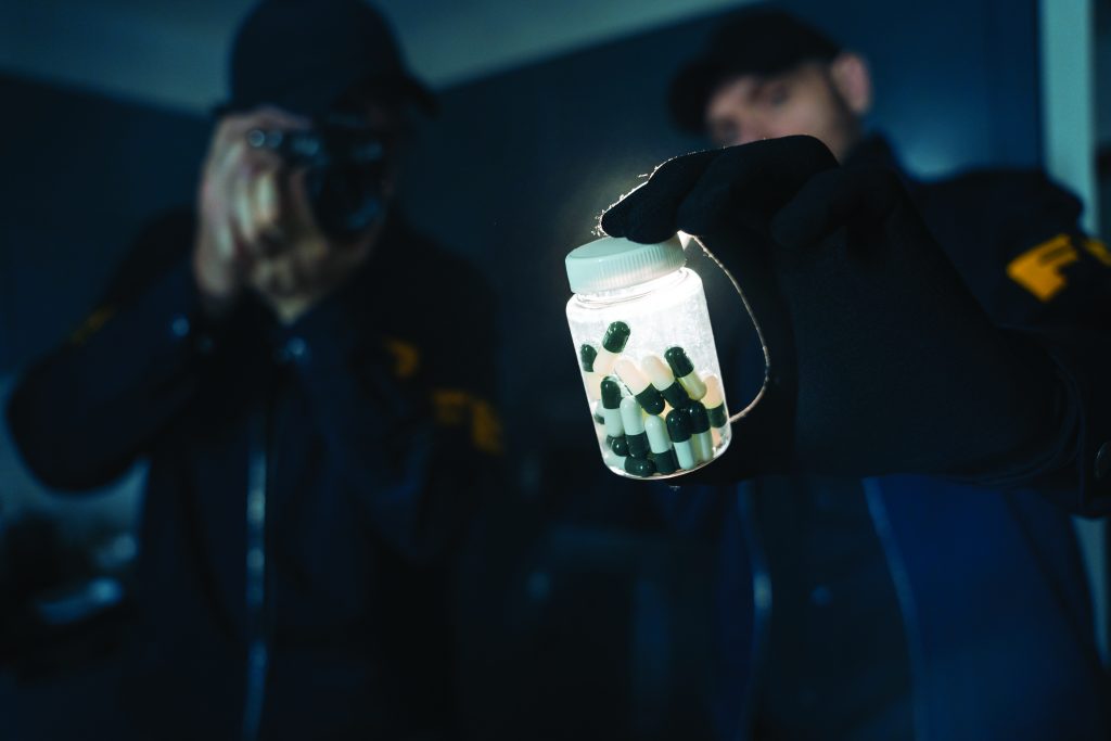 Two officers in dark uniforms; one holds a camera, the other displays clear plastic bottle with green and white capsules, documenting seized evidence