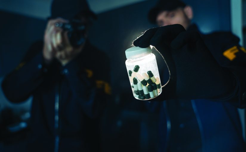 Two officers in dark uniforms; one holds a camera, the other displays clear plastic bottle with green and white capsules, documenting seized evidence