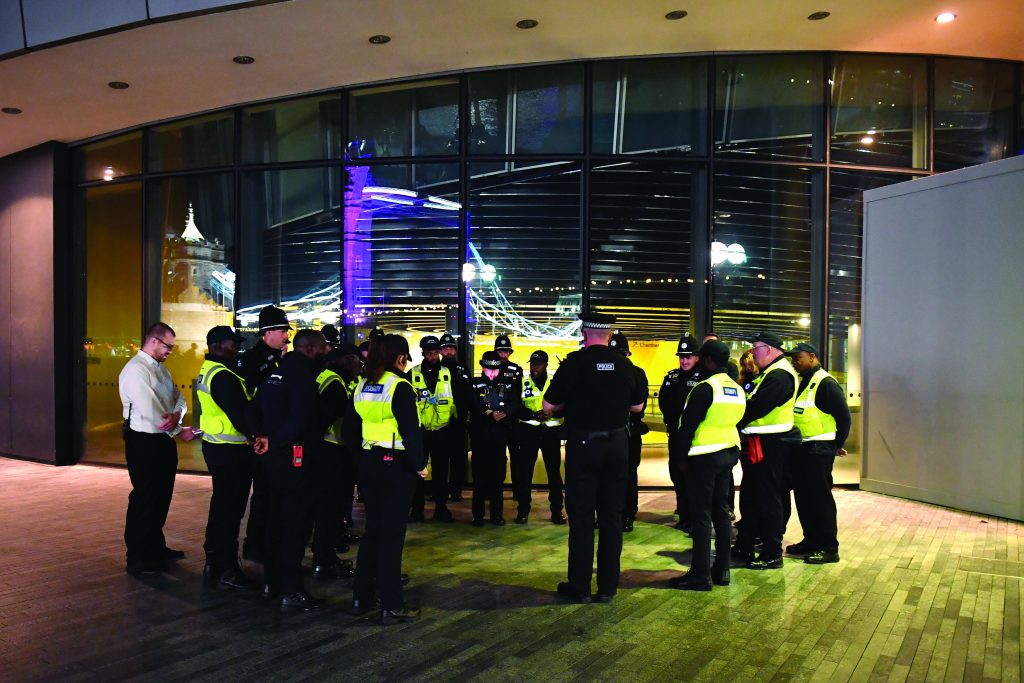 Uniformed police and security personnel in high‑visibility vests stand in a nighttime briefing outside a glass‑fronted building, with radios, helmets, city lights, and bridge reflections visible