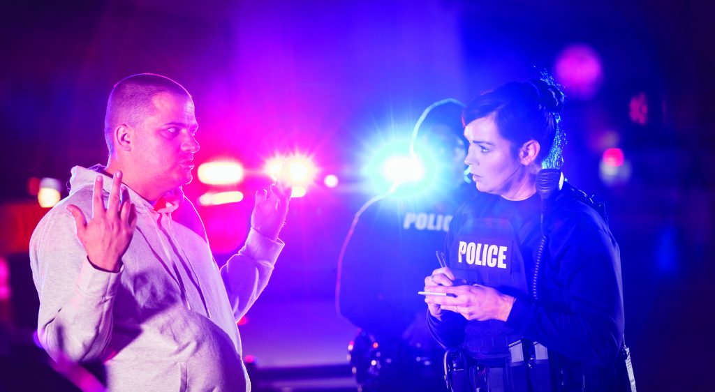 Two multiracial police officers working at night, taking a statement from a victim or witness to a crime. It is nighttime, and the emergency lights of a police car are visible in the background.  The focus is on the policewoman and the civilian in the foreground. The other officer is an African-American man.