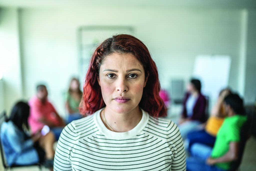 Person in striped shirt standing in focus with a group seated in a circle in the background, inside a bright room with whiteboard and chairs.