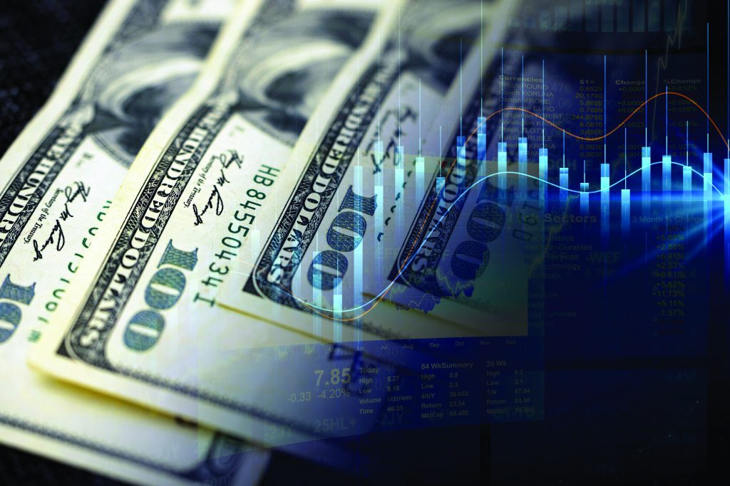 The image shows several overlapping U.S. one hundred dollar bills arranged in a fan-like manner. Overlaid on the right side of the image is a transparent graphic of a financial chart with blue and red lines indicating stock market trends, along with various numerical data points and bar graphs. The background is dark, creating a contrast that highlights both the currency and the financial chart elements.