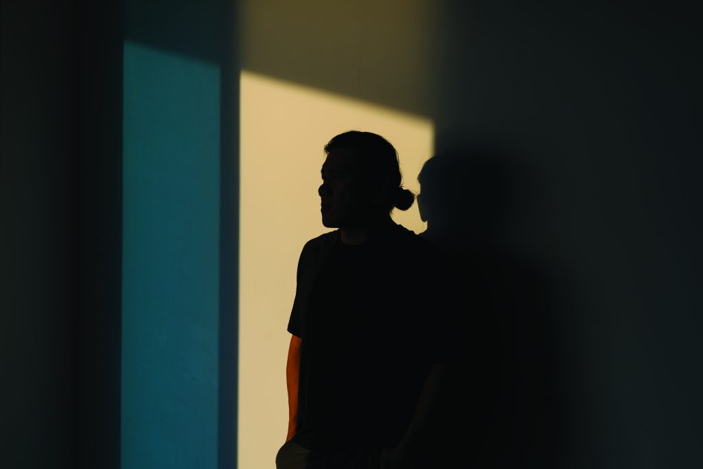 A dark silhouette stands against intersecting panels of light and shadow on a wall, with soft illumination creating sharp contrasts and a second faint outline
