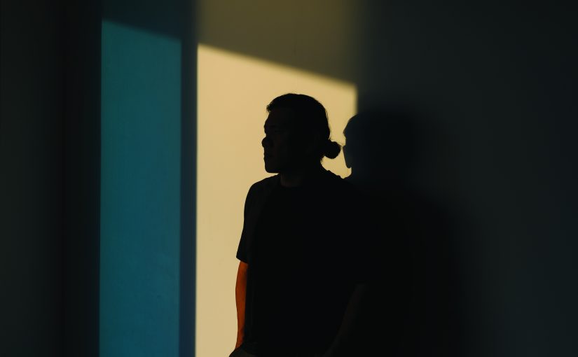 A dark silhouette stands against intersecting panels of light and shadow on a wall, with soft illumination creating sharp contrasts and a second faint outline