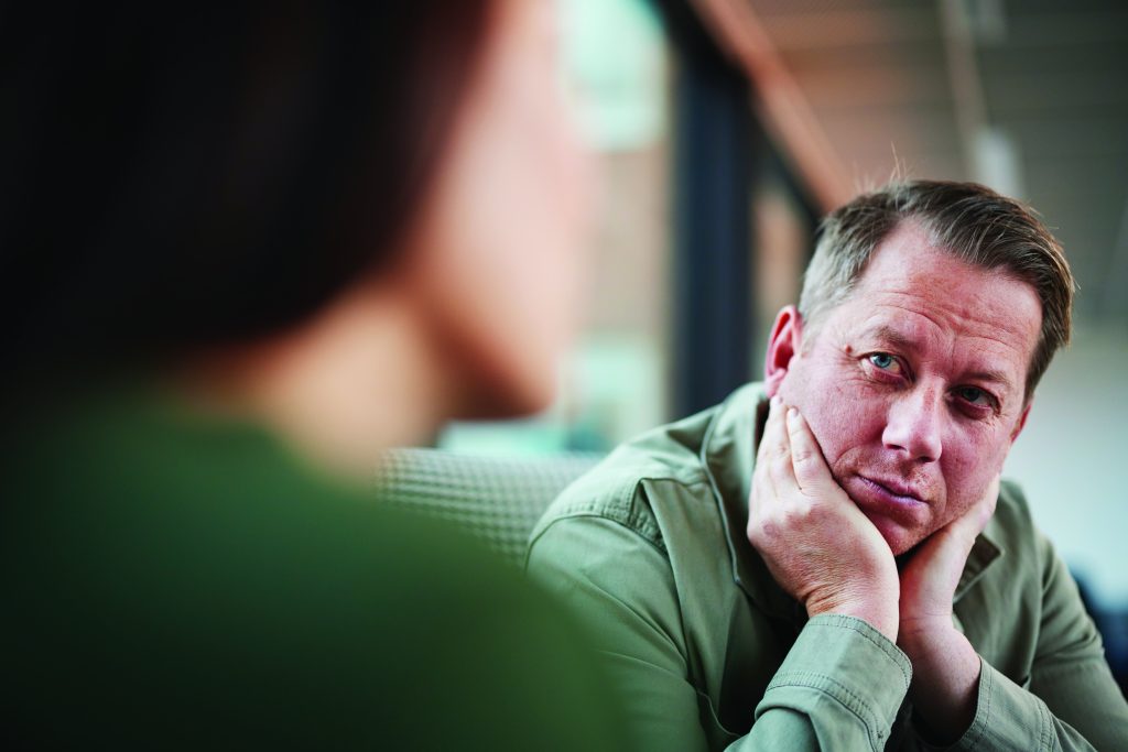 Two individuals seated across from each other in bright room with large windows; one wears green shirt with hands resting on face; indoor setting