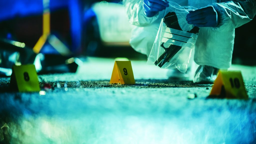 Forensic investigator in protective suit bags evidence near numbered crime-scene markers on pavement under blue lighting, with scattered debris visible