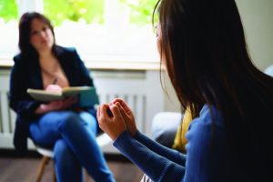 Close-up of person in blue sweater speaking during a counseling session, hands gesturing, with another individual seated holding an open notebook in bright room