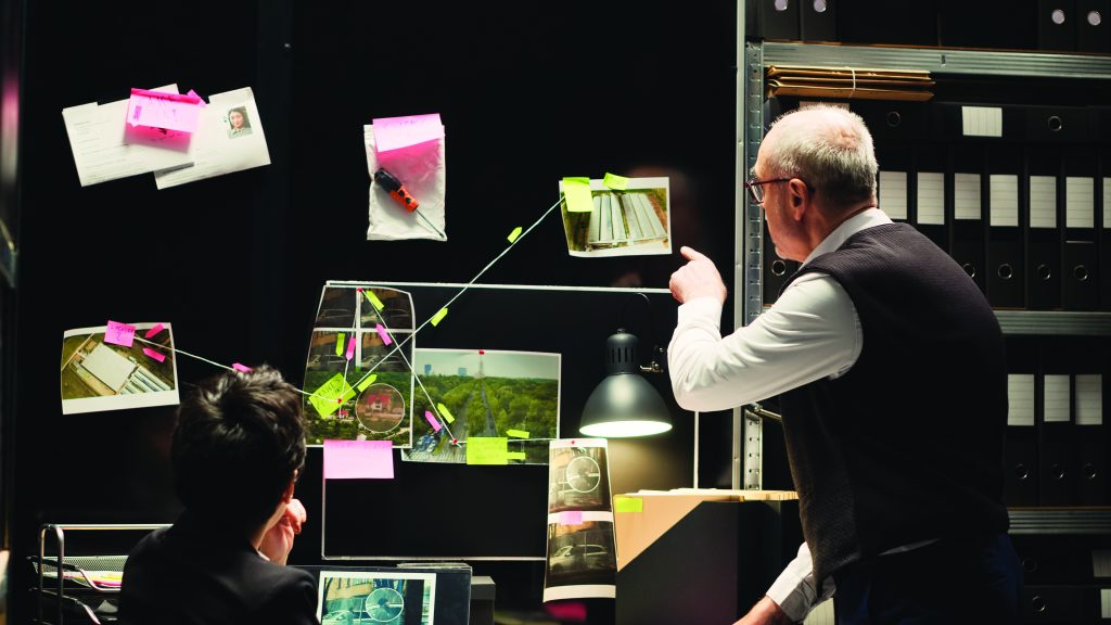 Investigator pointing at evidence board with photos, documents, and string connections under a desk lamp, while another person reviews case details on a laptop
