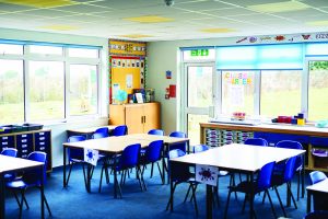 Bright classroom with blue chairs, rectangular tables, large windows, bulletin board decor, and 'Class Charter' poster on wall.