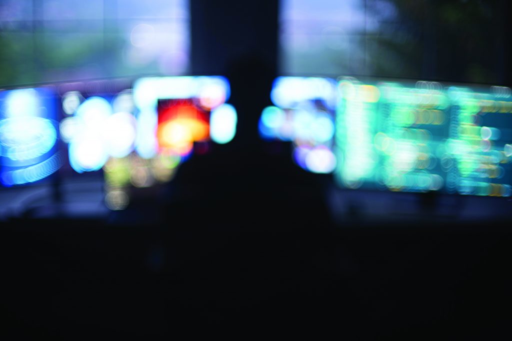 A silhouetted figure sits before multiple bright, blurred computer monitors displaying colorful data and graphics, creating a hazy, high‑tech workstation scene.