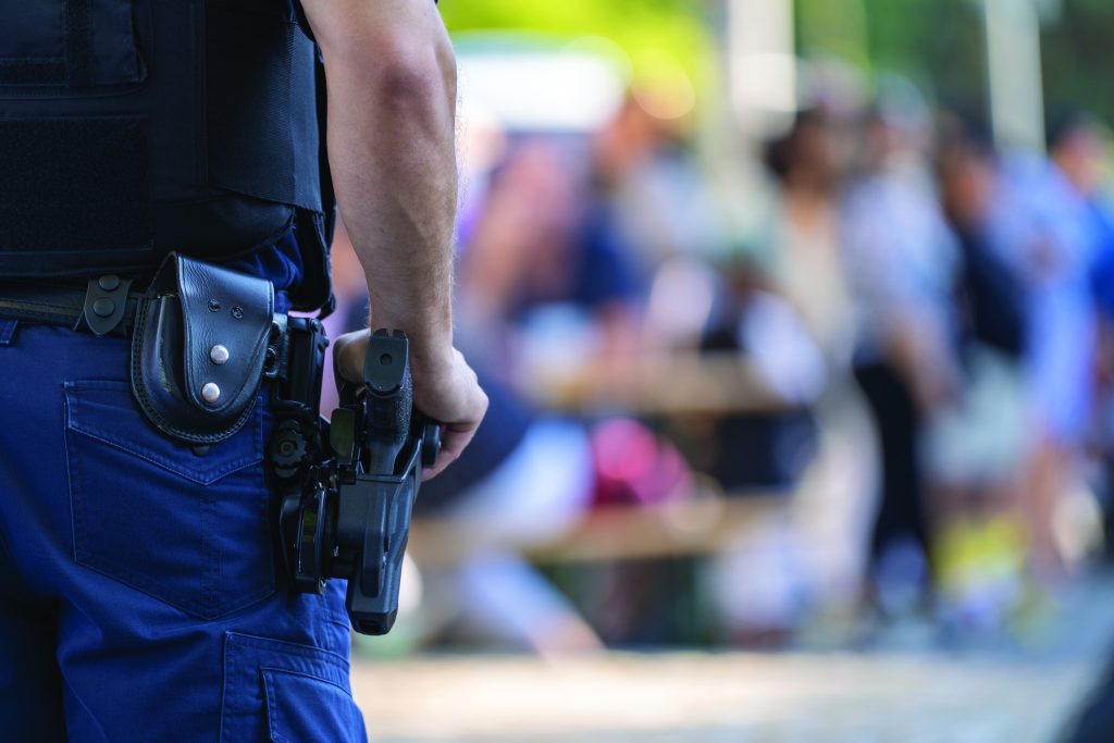 Uniformed law enforcement officer with holstered gun stands in focus; blurred crowd in background suggests presence at a public event or gathering