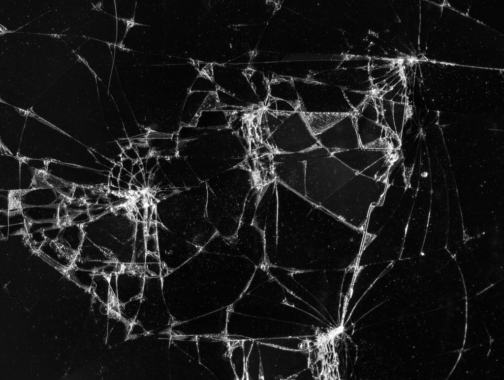 Close-up of shattered glass with complex, web-like cracks radiating outward on a dark background, highlighting impact patterns and fracture detail