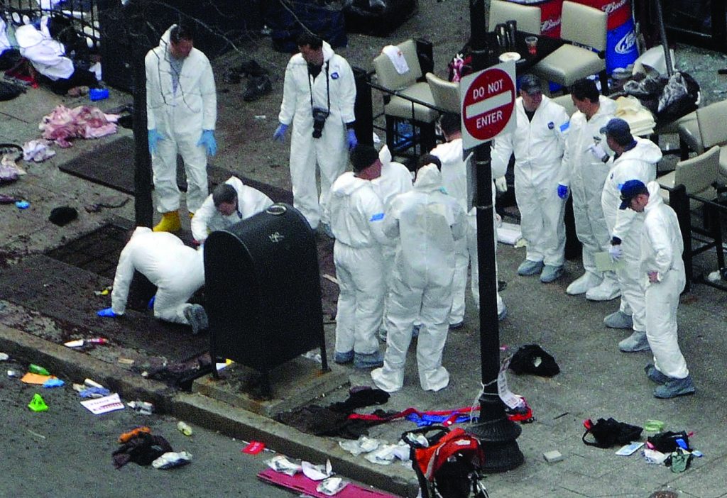 Forensic investigators in white suits and blue gloves examine debris on city sidewalk near 'Do Not Enter' sign and mailbox during daylight.