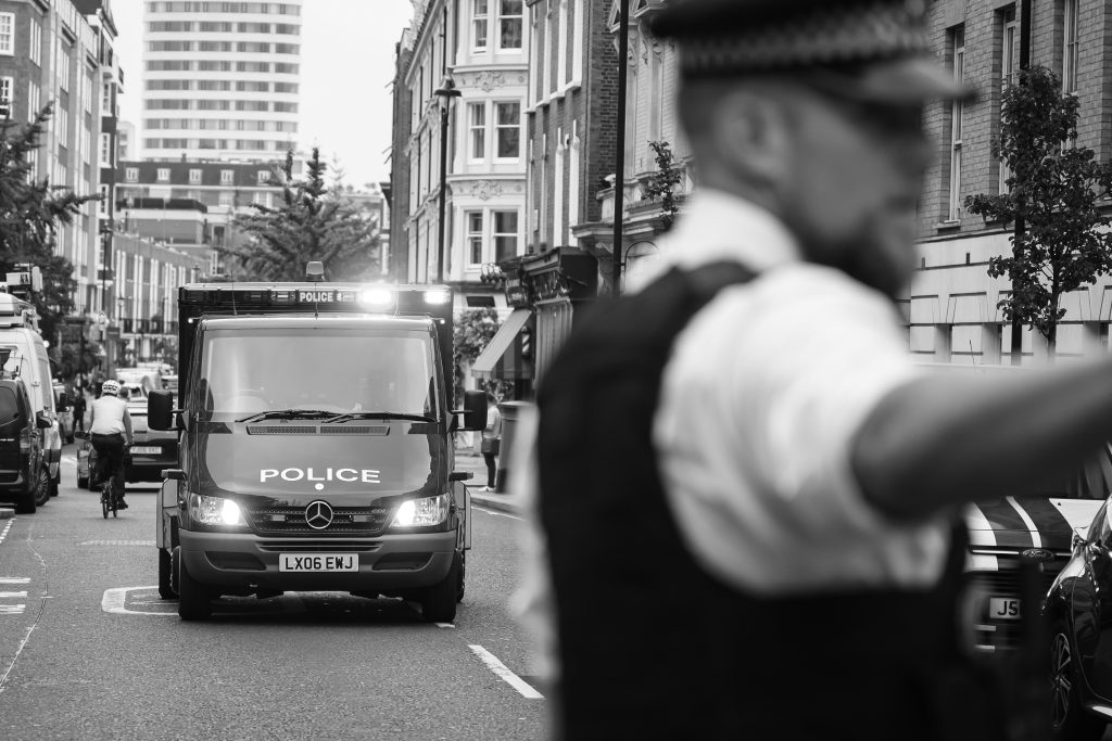 A police van with flashing blue lights moves down a city street lined with buildings as a uniformed officer in the foreground directs traffic and pedestrians