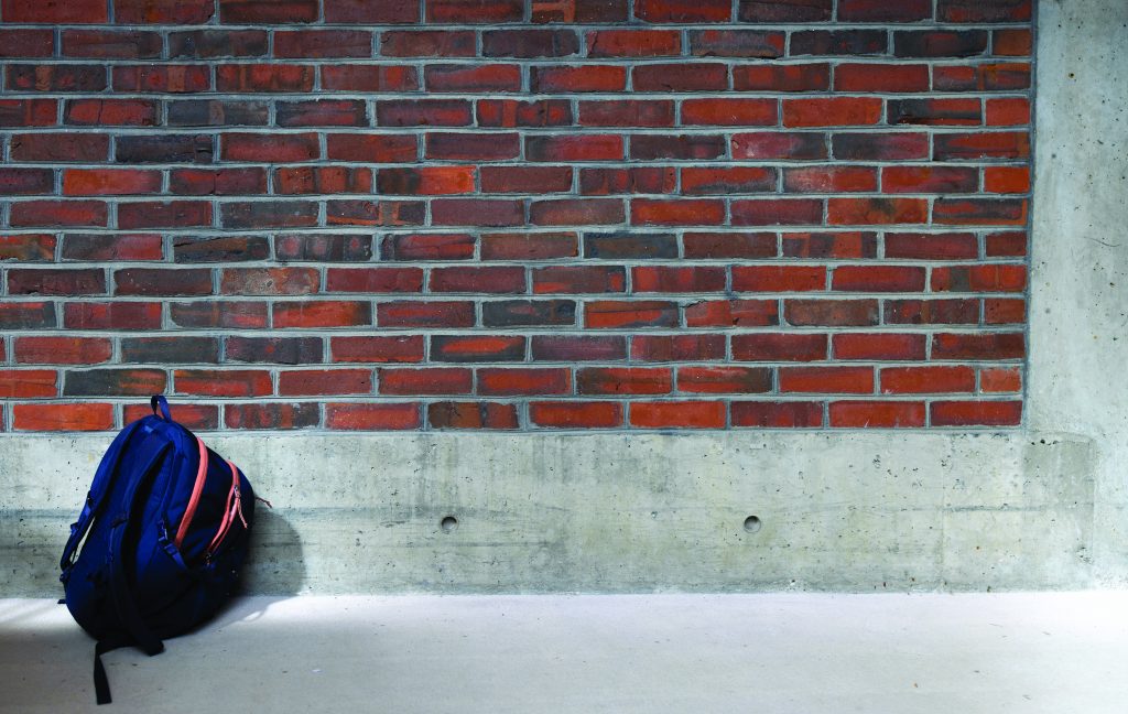 Blue and orange backpack leaning against red brick wall with concrete base; shadow cast on light floor in quiet, indoor setting.