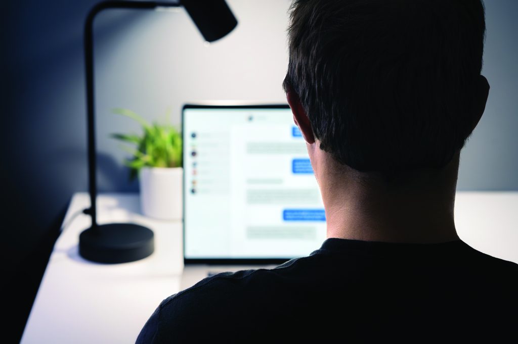 Person sitting at a desk views a laptop displaying a chat window, with a desk lamp and potted plant nearby, illuminated by soft indoor lighting