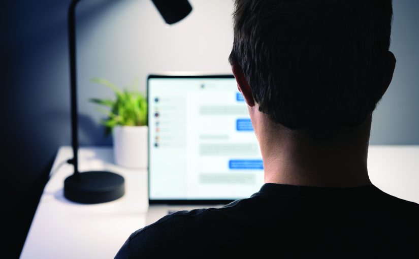 Person sitting at a desk views a laptop displaying a chat window, with a desk lamp and potted plant nearby, illuminated by soft indoor lighting