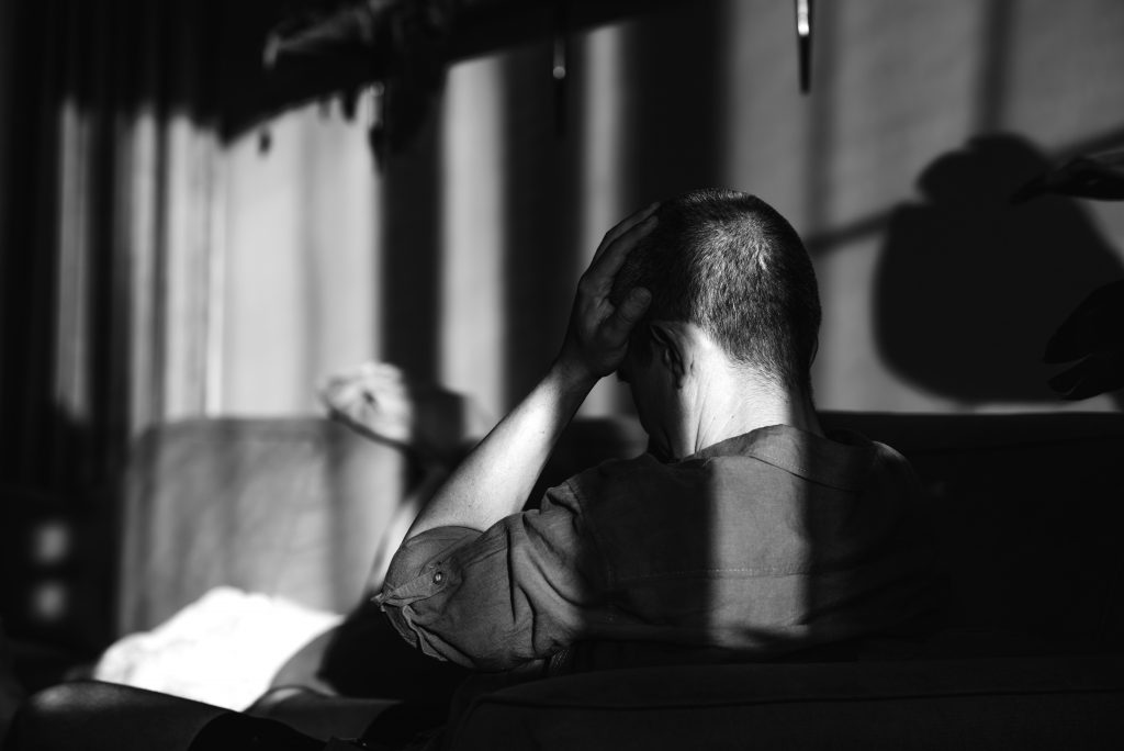 Black‑and‑white interior scene shows a seated individual from behind with one hand resting on the head, strong window light casting shadows across a chair, wall, and furnishings