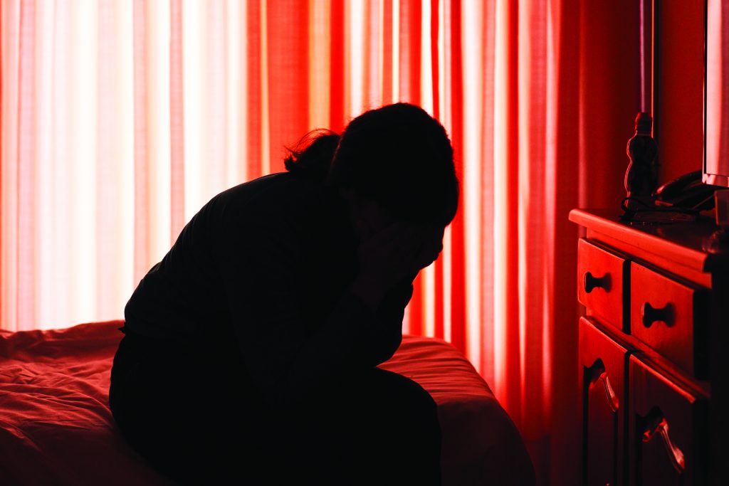 Silhouette of person seated on bed in dim room with red curtains, head in hands, beside dresser with scattered items, evoking stress or crisis setting