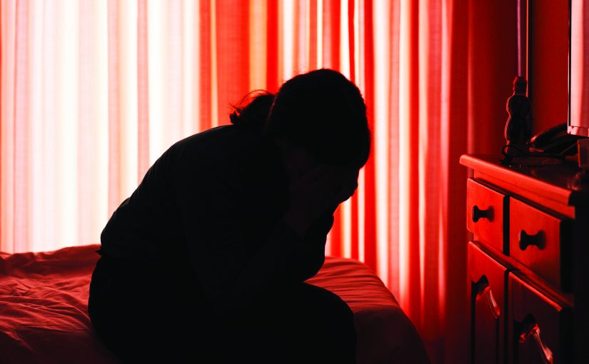Silhouette of person seated on bed in dim room with red curtains, head in hands, beside dresser with scattered items, evoking stress or crisis setting