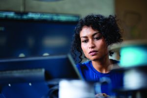 Individual with curly hair at multi-monitor workstation, surrounded by tech equipment in blurred office, emphasizing digital operations and analysis