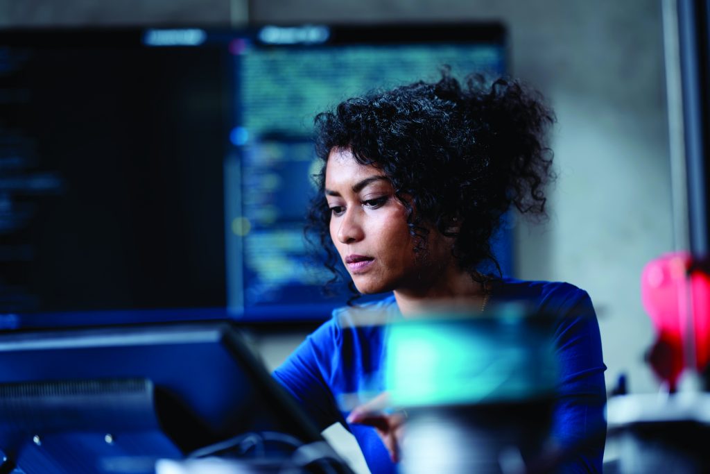 Person in a tech workspace sits near monitors displaying code, reaching toward equipment as screens glow behind them in a dim, modern cybersecurity setting