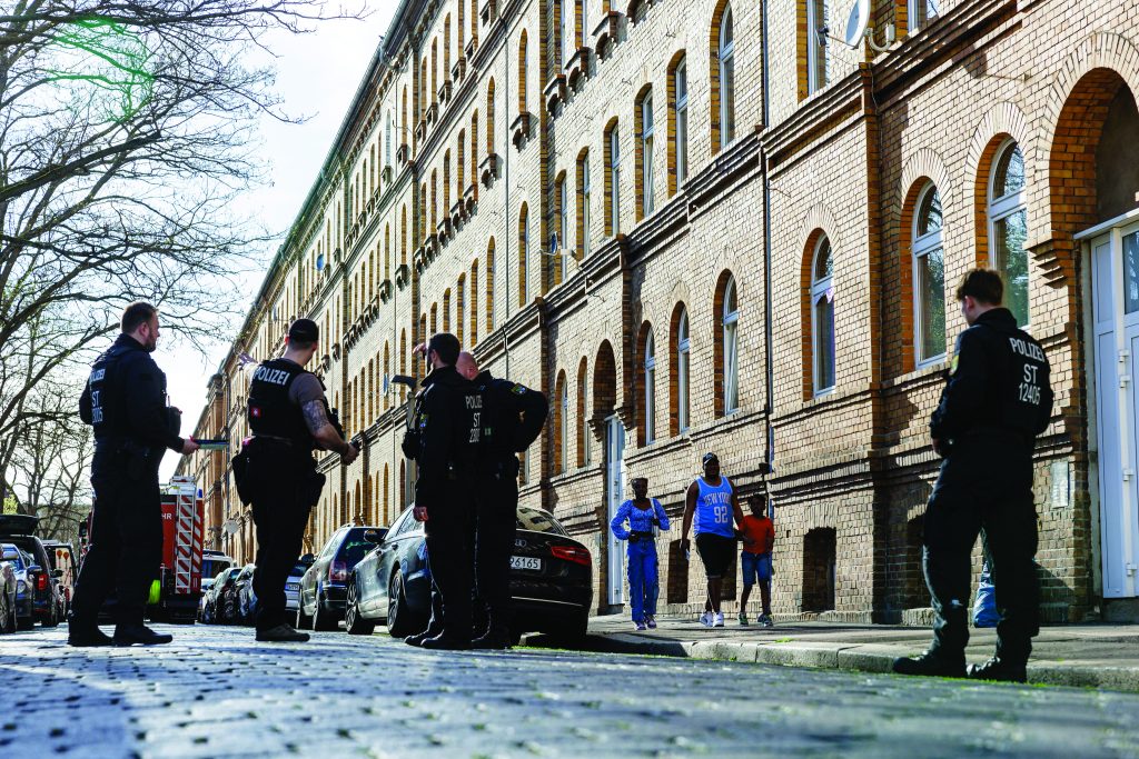 Four uniformed officers marked ‘POLIZEI’ stand along a cobblestone street beside parked vehicles and a long brick apartment building, with bare trees, pedestrians, sidewalks, windows, and architectural details visible in daylight