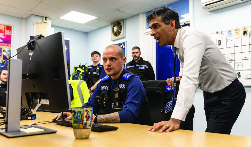 Police personnel work at desks in a busy office as one individual at a computer is assisted by another standing nearby, with uniforms, monitors, and wall boards visible
