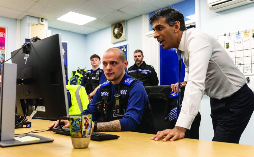 Police personnel work at desks in a busy office as one individual at a computer is assisted by another standing nearby, with uniforms, monitors, and wall boards visible