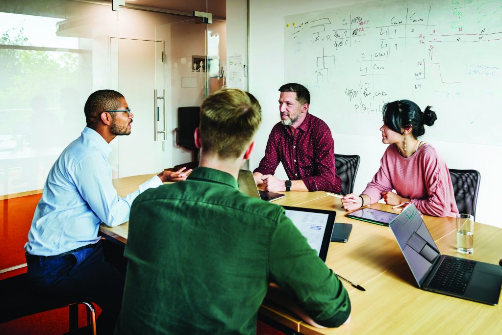 Four people sit around a conference table with laptops and tablets, discussing notes beside a whiteboard filled with diagrams, charts, and written annotations