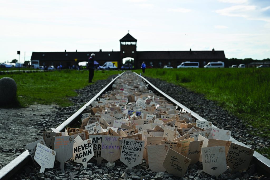 Rail tracks stretch toward a brick gate at Auschwitz, lined with handwritten memorial placards, visitors, parked vans, grass, gravel, and overcast sky