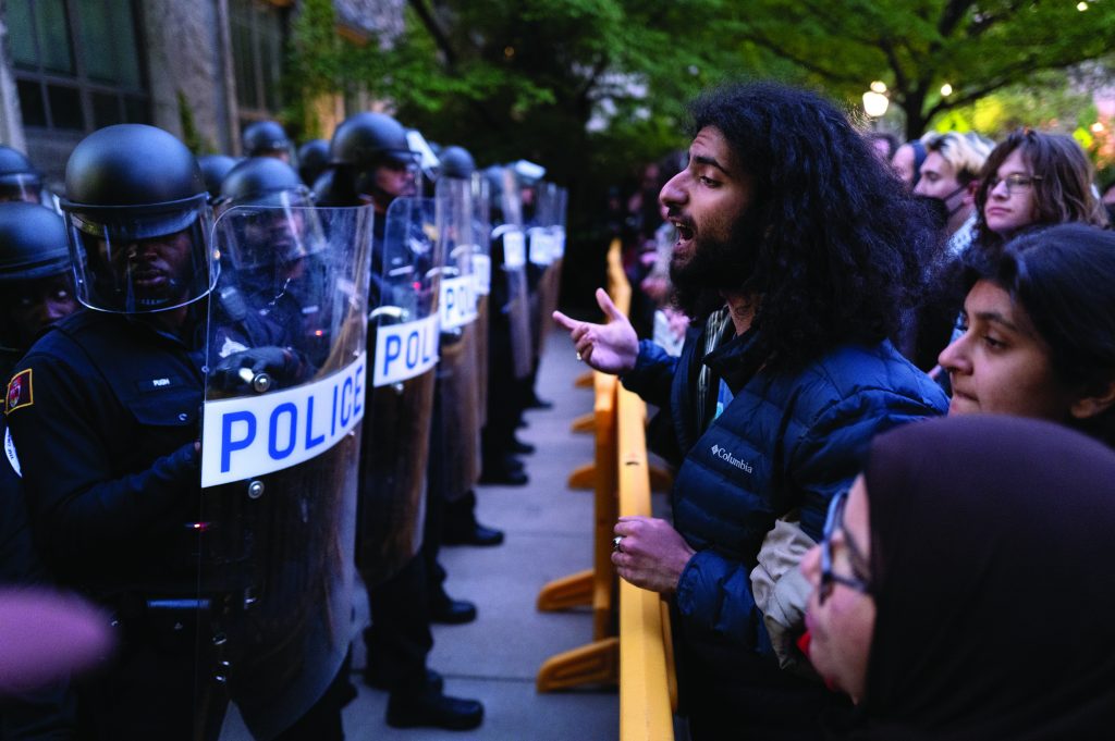 Line of officers in riot gear with 'POLICE' shields face protesters behind yellow barricade; one protester gestures passionately.