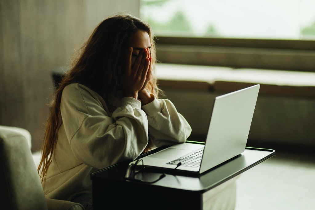 A person sits at a small table using an open laptop, resting both hands against the head, with a chair, window light, tabletop, and eyeglasses visible nearby