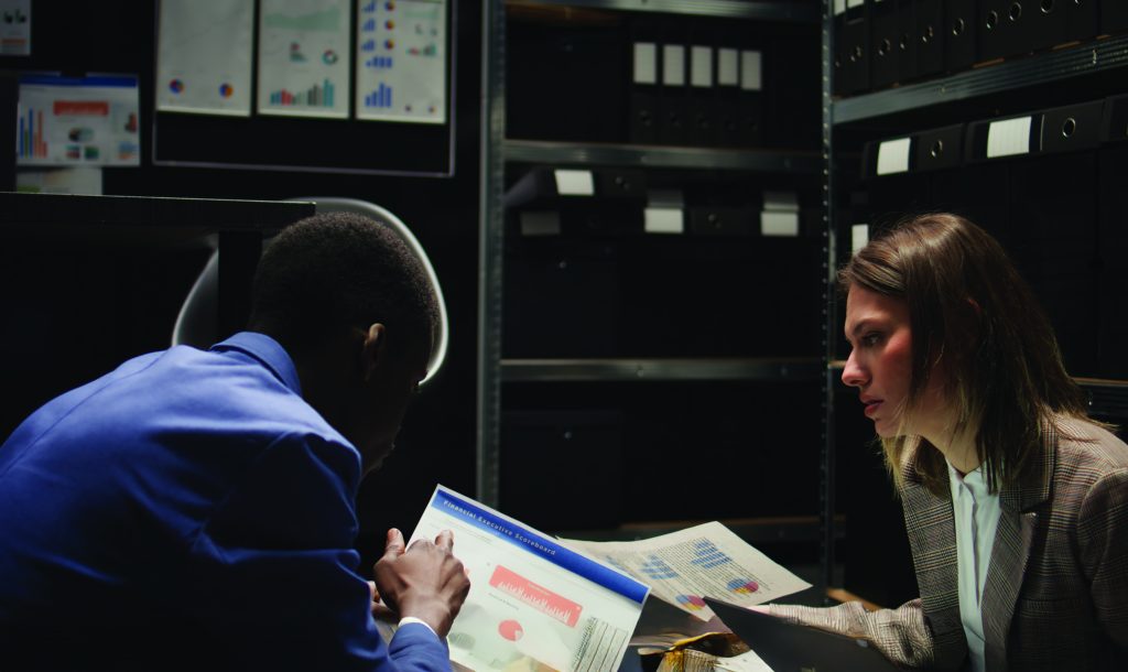 Two individuals review printed charts and graphs at a desk in a dimly lit office with shelves of black binders and wall-mounted data charts in the background