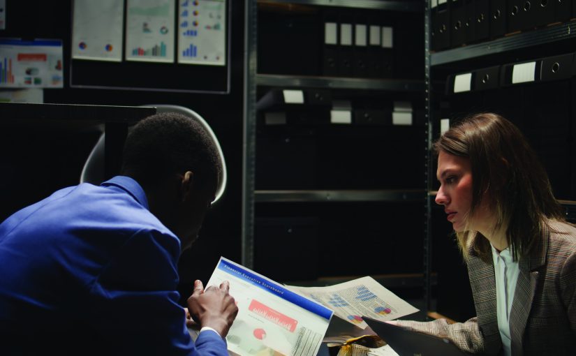 Two individuals review printed charts and graphs at a desk in a dimly lit office with shelves of black binders and wall-mounted data charts in the background