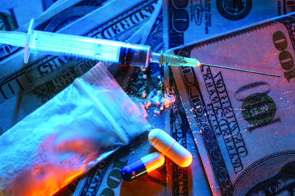Syringe, small bag of white powder, two capsules, and scattered $100 bills under dramatic blue-orange lighting—depicting illicit drug trade elements