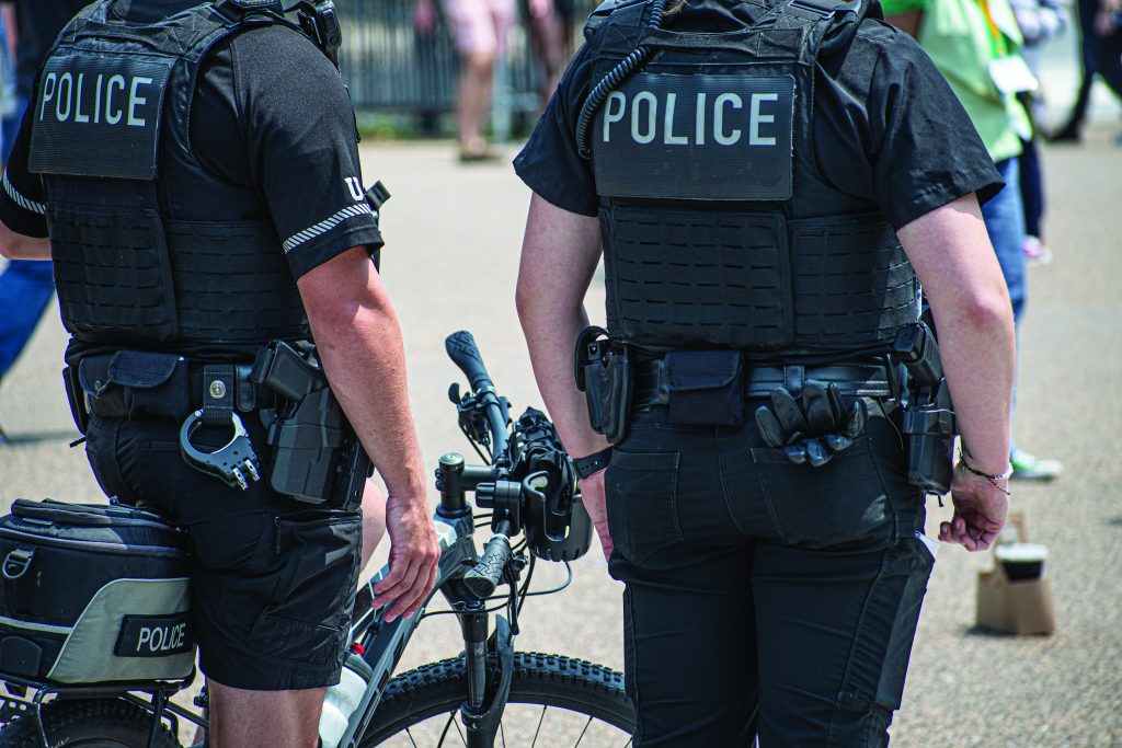 The image shows two law enforcement officers from behind, both wearing black uniforms with the word "POLICE" in white letters on their back. They are standing next to a police bicycle, which has a small storage bag attached to it labeled "POLICE." Both officers have various equipment attached to their belts, including handcuffs and radios. The officer on the left is holding onto the bicycle's handlebars. In the background, there are blurred figures of people walking and some indistinct objects.