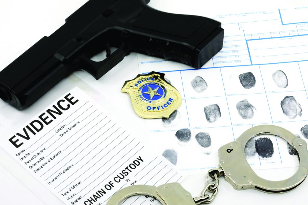 Black handgun, police badge, evidence bag with chain-of-custody form, fingerprint cards, and metal handcuffs displayed on flat surface.