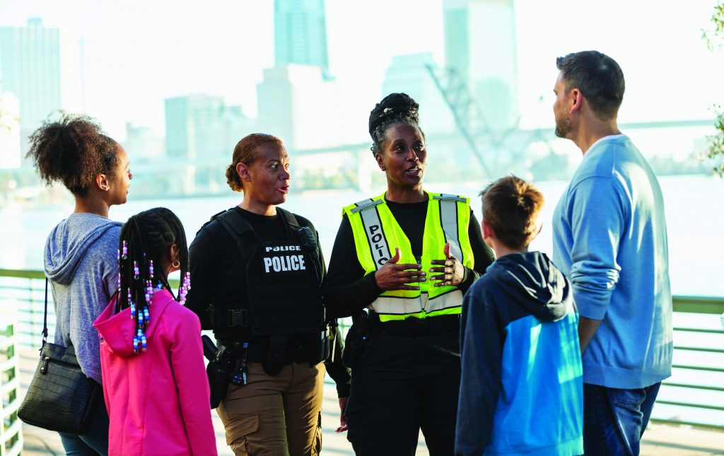 Two multiracial policewomen, in their 40s and 50s, conversing on a city waterfront with two families, getting to know people in the local community.