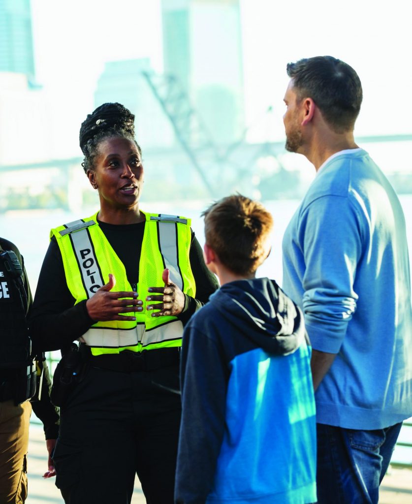 Two officers—one in black uniform, one in yellow vest—speak with man, woman, and two children on riverside walkway; city buildings in background