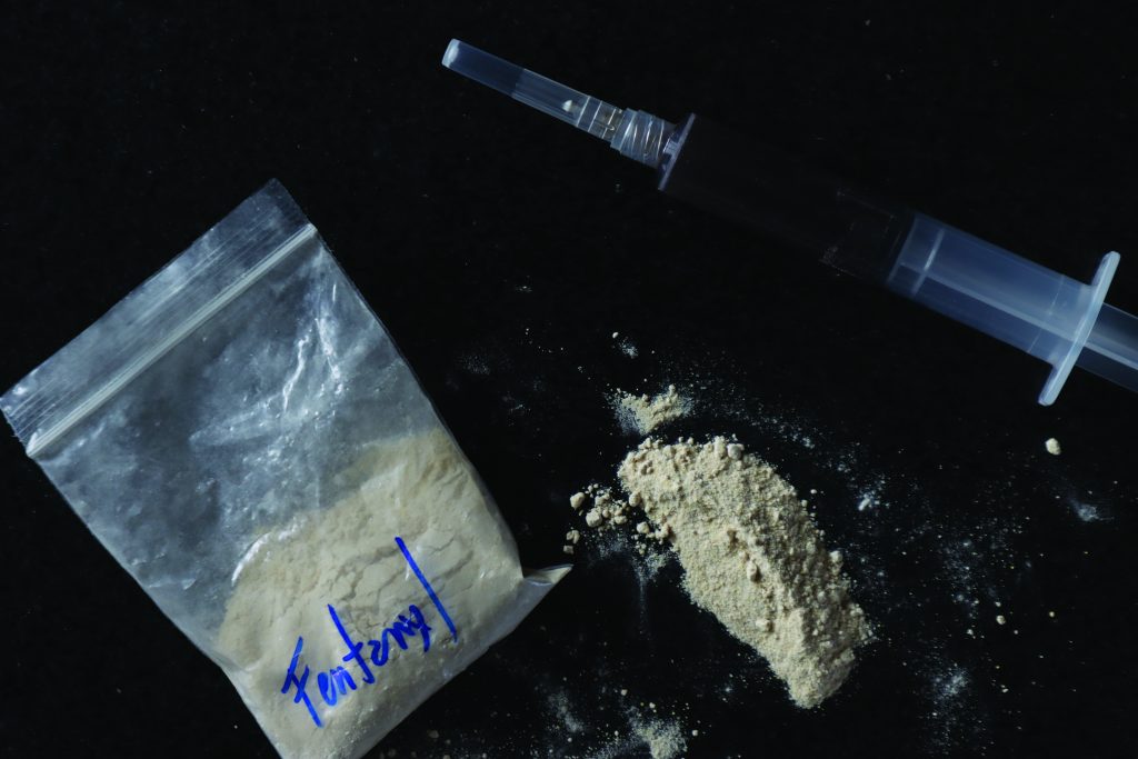 Clear bag labeled 'Fentanyl' in blue ink beside beige powder pile and capped syringe, all placed on a dark surface, highlighting drug evidence
