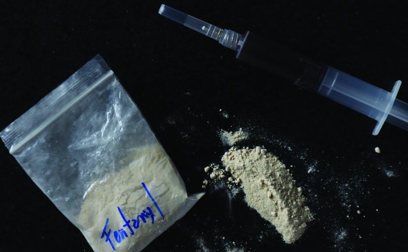 Clear bag labeled 'Fentanyl' in blue ink beside beige powder pile and capped syringe, all placed on a dark surface, highlighting drug evidence