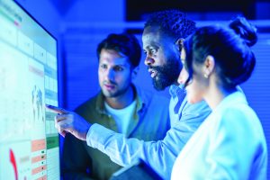 Three people review a large digital touchscreen displaying data panels and a world map in a blue-lit operations workspace, one pointing at the screen