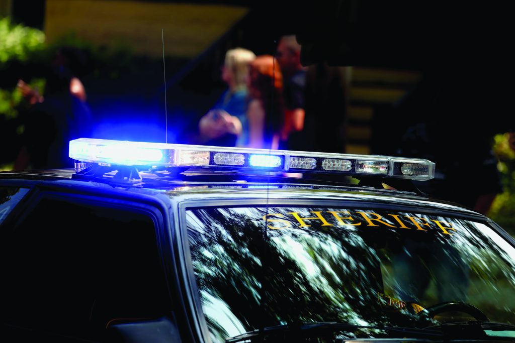 Sheriff patrol car with flashing blue-white lights parked outdoors; 'SHERIFF' marked on windshield; blurred figures in background suggest active scene.