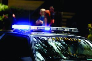 Sheriff patrol car with flashing blue-white lights parked outdoors; 'SHERIFF' marked on windshield; blurred figures in background suggest active scene.