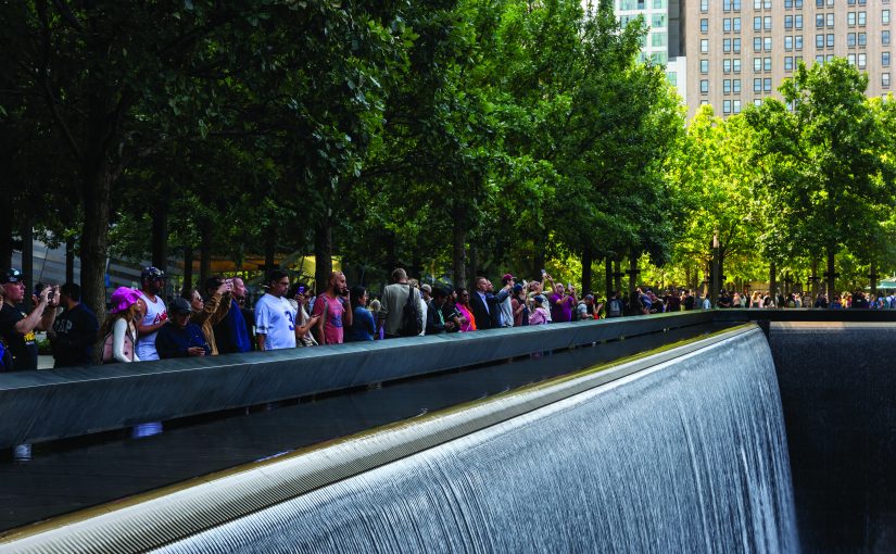 Visitors line the edge of the National September 11 Memorial reflecting pool, observing cascading waterfalls and engraved parapets beneath rows of trees, with surrounding office towers, paved plaza walkways, and dense crowds stretching along the memorial perimeter