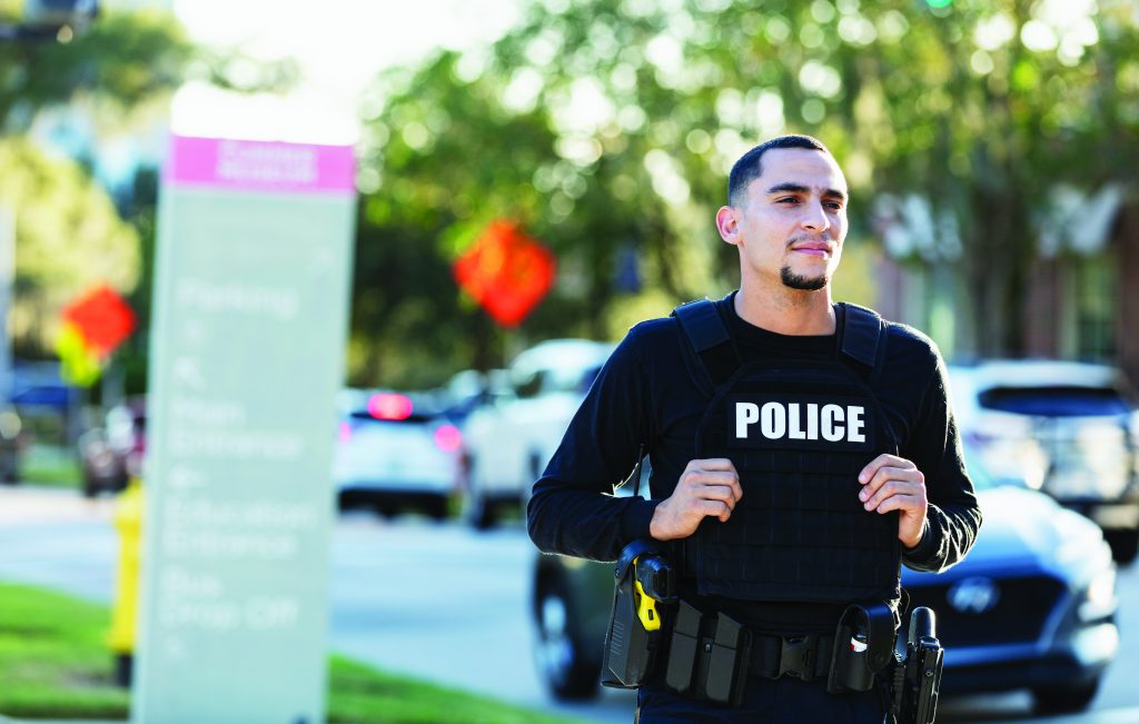 The image shows a police officer jogging on a street. The officer is wearing a black tactical vest with the word "POLICE" in white letters across the chest. The officer has short hair and is looking straight ahead, but the face is blurred for privacy. In the background, there are several cars on the road and an out-of-focus signpost with various texts. There are also trees and some orange construction signs visible in the distance. The overall scene appears to be during daylight hours with natural lighting.