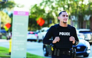 The image shows a police officer jogging on a street. The officer is wearing a black tactical vest with the word "POLICE" in white letters across the chest. The officer has short hair and is looking straight ahead, but the face is blurred for privacy. In the background, there are several cars on the road and an out-of-focus signpost with various texts. There are also trees and some orange construction signs visible in the distance. The overall scene appears to be during daylight hours with natural lighting.